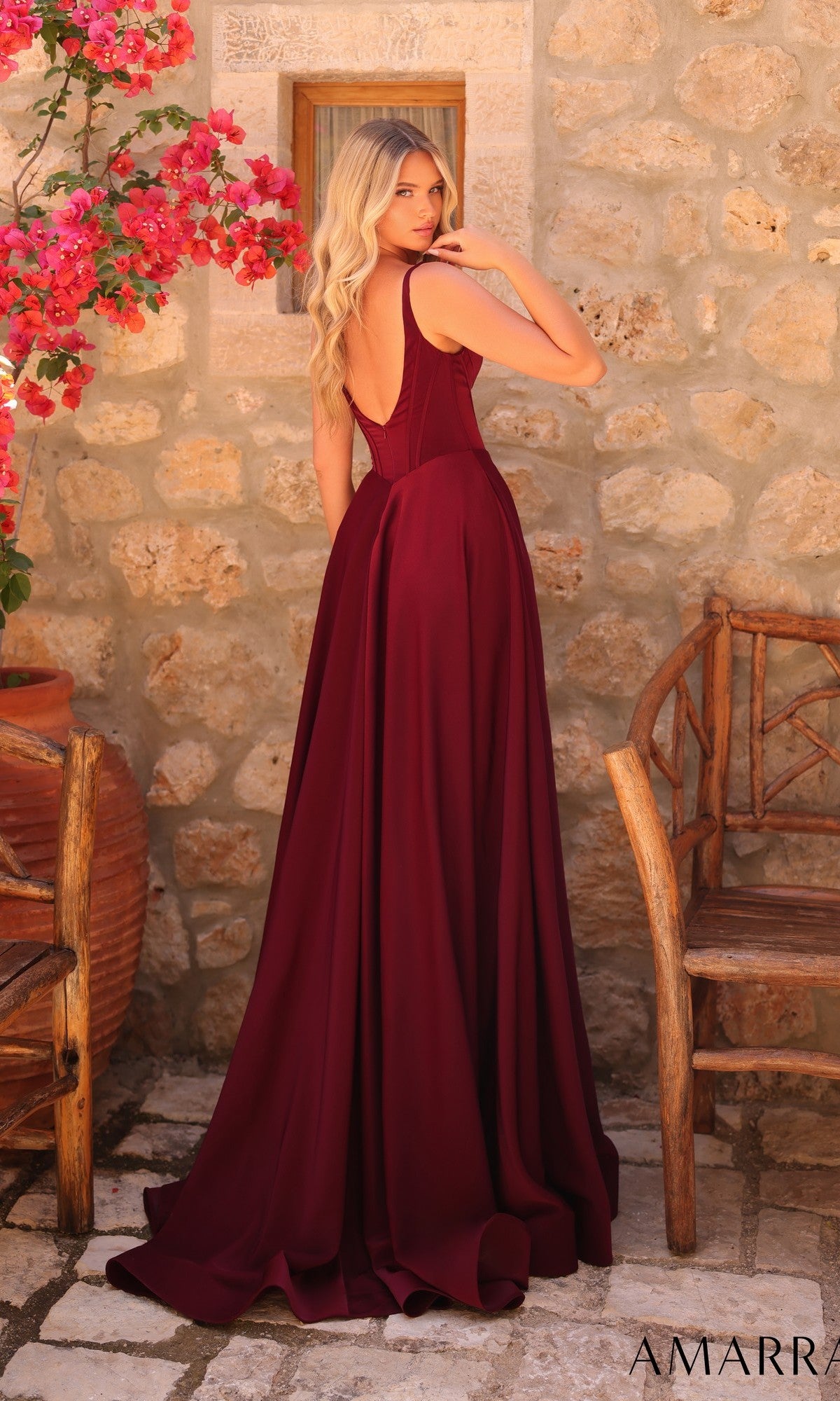 A woman with long blonde hair wears the Amarra 89519 long prom dress in burgundy, featuring a sleeveless design and low back, standing outdoors near a stone wall, wooden chair, and pink flowers.
